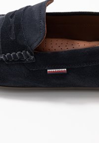 Mocassin en daim noir avec détails cousus, semelle intérieure en cuir marron perforée et petit logo en tissu rouge, blanc et bleu sur le côté.