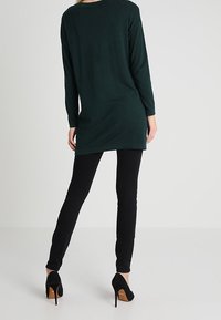 Tunica lunga verde scuro con maniche lunghe e scollatura rotonda, abbinata a pantaloni neri slim-fit e tacchi neri con un accento marrone sul tacco.