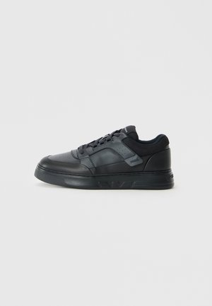 Zapatilla negra de caña baja con cordones en la parte delantera, parte superior de cuero perforado y cuello acolchado, mostrada sobre un fondo claro y simple.