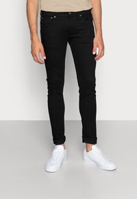 Jeans ajustados negros de mezclilla, con cinco bolsillos, cierre de botón y cremallera, y dobladillos enrollados, acompañados de zapatillas blancas.