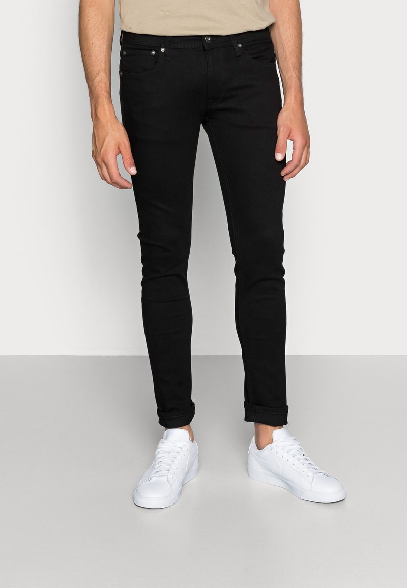 Jeans ajustados negros de mezclilla, con cinco bolsillos, cierre de botón y cremallera, y dobladillos enrollados, acompañados de zapatillas blancas.