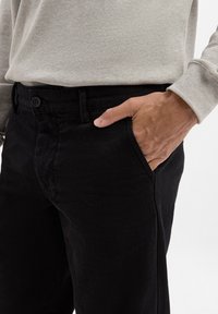 Jean noir en tissu texturé, avec une poche avant et une fermeture à bouton, associé à un haut à manches longues gris clair.