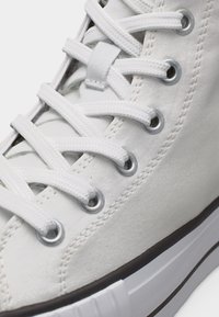 Artimo vaizdas į baltą aukštakulnį sportinį batelį su baltais raišteliais, prasuktais per metalines skylutes ir siūtu drobiniu viršumi.