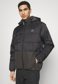 Black and dark green insulated puffer jacket with a zip front, adjustable hood, and side pockets. Features a logo on the chest.