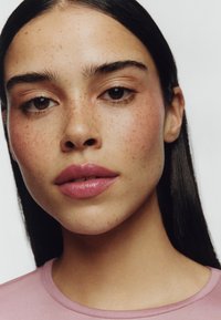 Portrait einer Person mit klarer Haut, Sommersprossen und vollen Lippen, die mit hellrosa Lippenstift betont sind. Sie hat glattes, dunkles Haar und trägt ein rosa Oberteil.