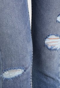 Vaqueros de mezclilla azul desgastados con grandes roturas irregulares en las rodillas, con bordes deshilachados y un patrón texturizado tejido.