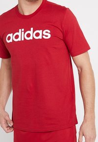 Muž v červeném tričku Adidas s bílým logem, záběr oříznutý na trup a paže na jednobarevném pozadí.