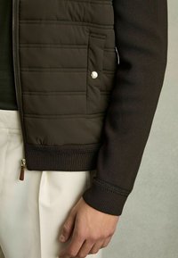 Jachetă păturată maro, cu mâneci și guler din tricot cu model în relief, având o palmă laterală cu buton cu clichet, talie din tricot detaliată și fermoar.