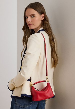 Pikkade pruunide juustega naine, kes kannab valget bleiserit tumesiniste ääriste ja kuldsete nuppudega, ning kannab väikest punast nahast õlakotti.