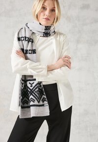Šedý a černý vzorovaný šátek vyrobený z měkké látky, s geometrickými designy. Modelka má na sobě bílé triko a černé kalhoty, s překříženýma rukama.