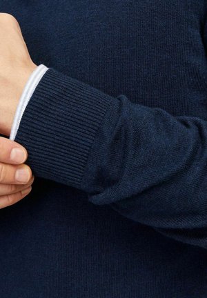 Jūrų mėlynos spalvos megztinis su raukšlėtais rankogaliais. Rankogalyje matoma plona balta juostelė, suteikianti kontrastą tamsiai medžiagai. Minkšta tekstūra.