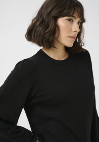 Maglione nero con scollatura rotonda classica e maniche puff testurizzate con motivi a diamante accentuati da piccole perline nere.