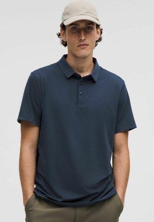 Homme portant une casquette de baseball beige, une chemise polo à manches courtes bleu marine et un pantalon kaki, se tenant avec les mains dans les poches.
