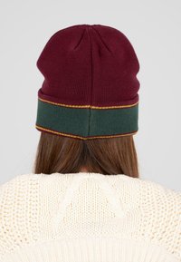 Gebreide beanie in bordeaux met een groene cuff en gele stiksels. Glad textuur met een afgerond ontwerp aan de bovenkant.