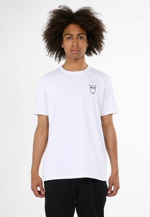 Jeune homme aux cheveux bouclés portant un t-shirt blanc uni avec un petit motif de chouette noire sur le côté gauche de la poitrine et un pantalon noir.