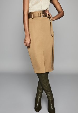 Mujer con falda beige hasta la rodilla con un cinturón ancho marrón, combinada con botas altas de color verde oliva, de pie frente a un fondo gris liso.