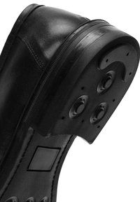 Chaussure en cuir noir avec une semelle en caoutchouc texturé, comportant quatre clous ronds et une pièce rectangulaire sur le côté. Fini lisse dans l'ensemble.