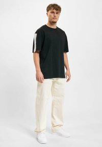 Juodas marškinėliai trumpomis rankovėmis, su baltais akcentais ant pečių, derinami su grietinėlės spalvos kelnėmis ir baltais sportbačiais.