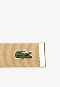 Ceinture en tissu beige tissé avec un logo crocodile vert brodé près de la fermeture métallique argentée.