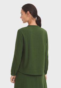 Haut vert à manches longues avec un col, fabriqué en tissu doux, présentant une texture lisse et des poignets boutonnés, vu de dos.