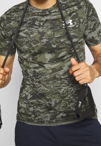 Zelené maskáčové tričko vyrobené z měkké látky. Má krátké rukávy, kulatý výstřih a bílé logo na levé straně hrudi.