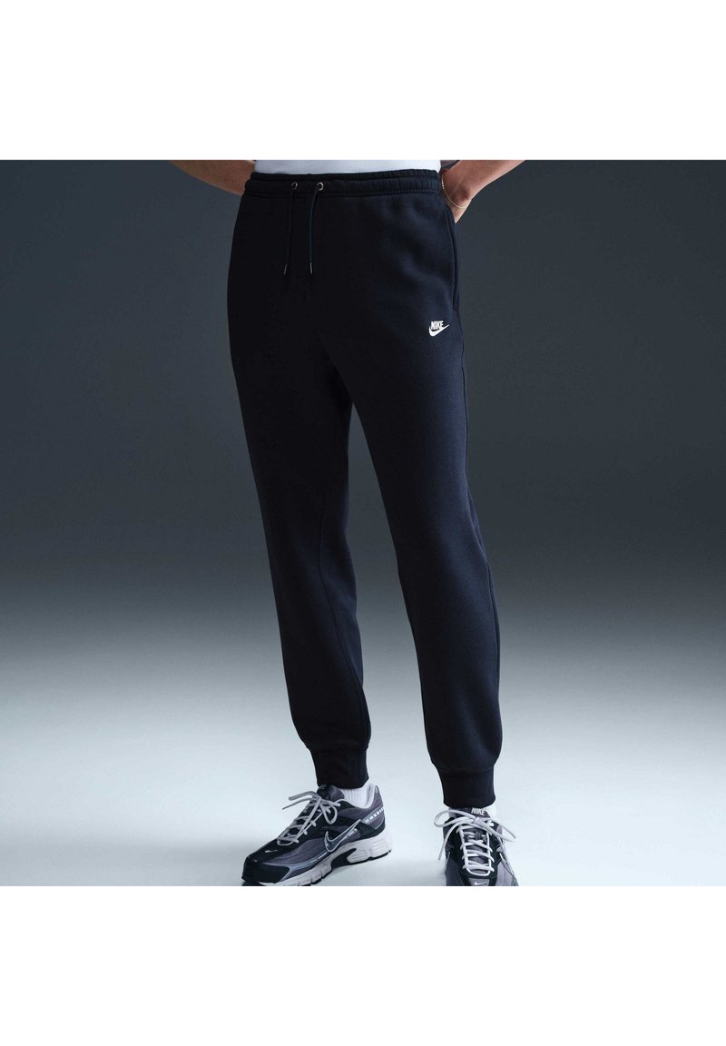 Persona che indossa pantaloni della tuta Nike neri e sneaker Nike nere e bianche, in piedi con le mani in tasca su uno sfondo scuro e uniforme.