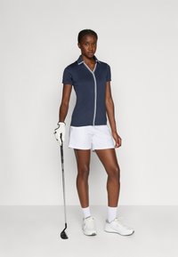 Námořnické polo tričko s krátkým rukávem a bílými detaily, spárované s bílými šortkami. Hráč drží golfovou hůl, má na sobě rukavici a bílé sportovní boty.