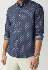 Man in een blauw overhemd met een klein rood en wit bloemenpatroon, crèmekleurige broek en een polshorloge met een bruine leren band.