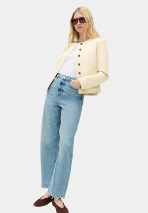 Kvinde iført oversized beige jakke med brune knapper, hvid top, lyseblå jeans, brune flade sko og solbriller, stående med den ene hånd i lommen.
