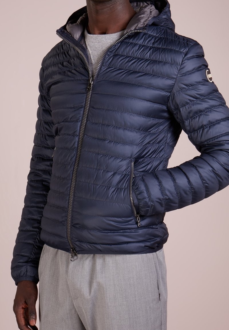 Marinblå pufferjacka med dragkedja, huva och sidofickor. Kvadrierad textur med en slät finish, med en kontrasterande innerfoder.