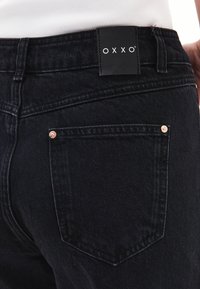 Svarta denimjeans med kopparnitar, med en närbild av bakfickan och en märkesetikett i svart på midjan.