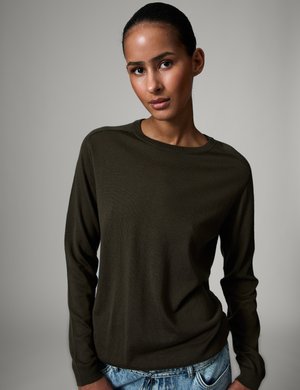 Žena s glatko počešljanom kosom, nosi tamnozeleno majicu dugih rukava i svijetloplave traperice, stoji ispred neutralne sive pozadine.