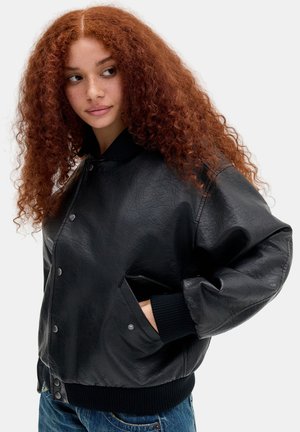 Jeune femme aux longs cheveux roux bouclés portant une veste en cuir noire avec des boutons et un jean bleu, regardant sur le côté avec la main dans la poche.