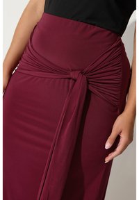 Vineröd kjol med knuten midja och rynkad struktur. Tillverkad av stretchigt material, slät yta, designad för en figurnära silhuett.