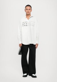 Weißes Hemd mit Knopfverschluss und gestreifter Textur, ausgestattet mit numerischen Grafiken auf der Vorderseite. Kombiniert mit weiten schwarzen Hosen und glänzenden schwarzen Schuhen.