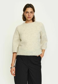 Hellbeige Strickpullover mit einem runden Ausschnitt, Dreiviertelärmeln und einer weichen, strukturierten Oberfläche. Kombiniert mit schwarzen Hosen.