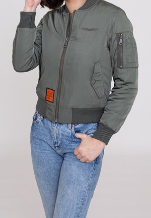Person wearing olive green bomber jacket with zip and pockets, paired with high-waisted blue jeans, hand in pocket, touching hair.