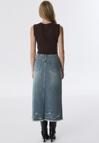 Braunes ärmelloses Oberteil, kombiniert mit einem langen, hellblauen Jeansrock mit ausgefranstem Saum und abgetragenen Details. Schwarze Ankle Boots runden den Look ab.
