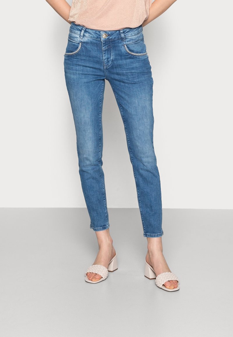 Femme portant un jean bleu slim taille mi-haute et des sandales beiges à talons avec des brides tressées, debout devant un fond clair uni.