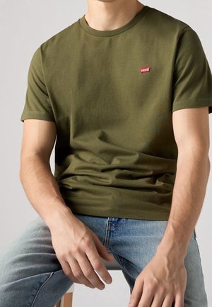 Man draagt een olijfgroen Levi's t-shirt en lichtblauwe spijkerbroek, zittend met handen op de dijen tegen een effen achtergrond.