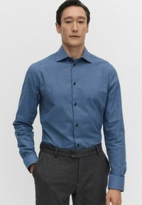 Niebieska koszula zapinana na guziki, wykonana z teksturowanej tkaniny, z kołnierzykiem, długimi rękawami i ciemnymi guzikami. Połączona z szarymi spodniami wizytowymi.