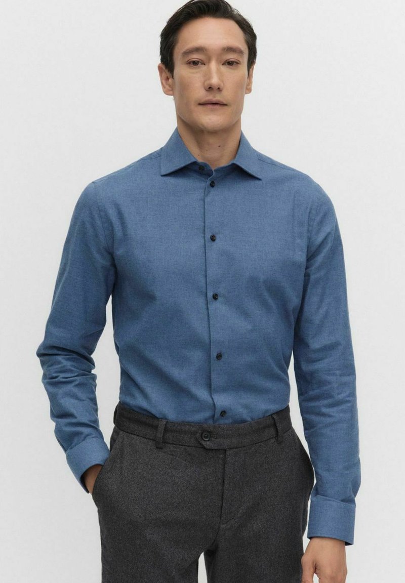 Niebieska koszula zapinana na guziki, wykonana z teksturowanej tkaniny, z kołnierzykiem, długimi rękawami i ciemnymi guzikami. Połączona z szarymi spodniami wizytowymi.