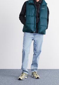 Grüne Puffweste über einem schwarzen Hoodie, hellblaue Jeans und beige Sneakers mit schwarzen und grünen Akzenten. Texturiertes Material mit einem Reißverschluss.