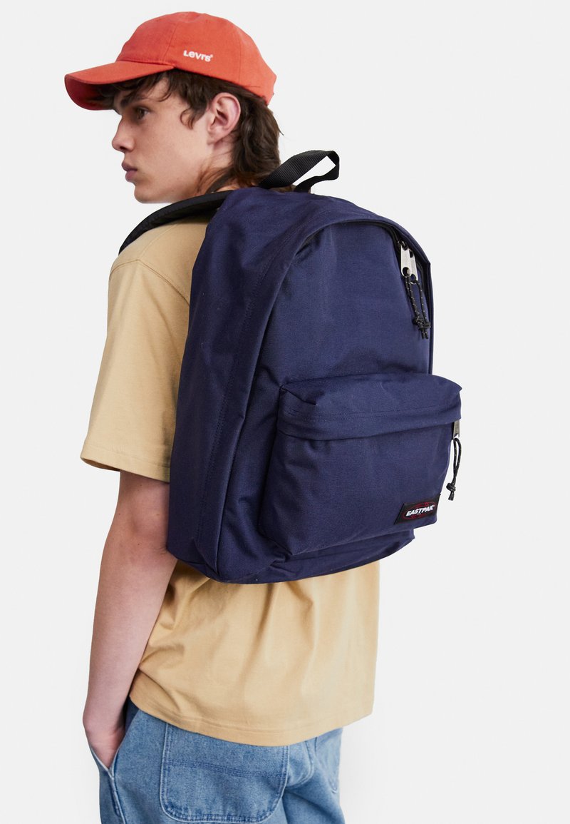 Navy fabric backpack with padded straps, front zip pocket, and logo tag. Worn over a beige shirt and paired with light blue jeans.