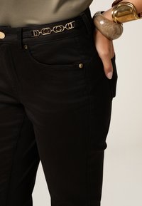 Los pantalones vaqueros de cintura alta en negro cuentan con herrajes dorados, bolsillos laterales y un cinturón con eslabones circulares. Tienen una textura suave y un corte entallado.