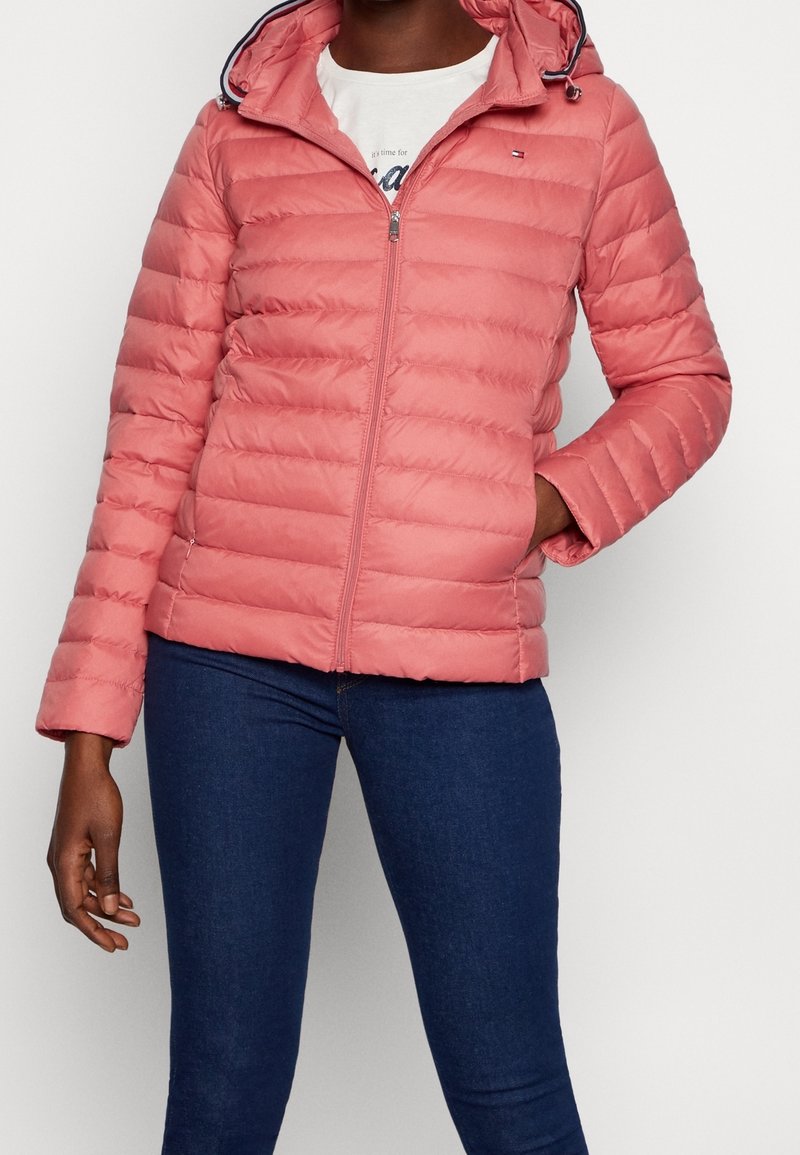 Femme portant une doudoune rose corail avec une capuche, fermée par une fermeture éclair, associée à un jean skinny bleu foncé, main dans la poche.