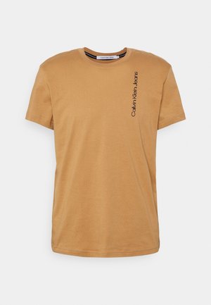 Mężczyzna w beżowej koszulce Calvin Klein Jeans z krótkim rękawem i niebieskich jeansach na jednolitym tle.