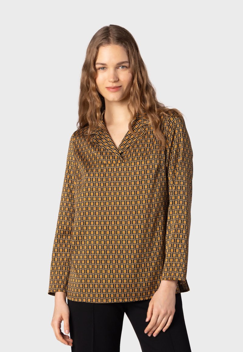 Femme aux longs cheveux ondulés porte une chemise marron à manches longues avec un motif carré noir et un pantalon noir, debout devant un fond uni.