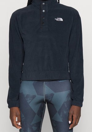 Personne portant un pull North Face noir à manches longues et des leggings de sport bleu foncé à motifs, debout les bras détendus le long du corps.