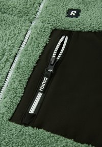 Tessuto di felpa verde con un pannello nero. Presenta due zip bianche e un piccolo etichetta con logo. Texture morbida; design casual e funzionale.